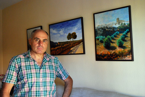 El pintor en el salón de su casa rodeado de sus óleos de paisajes alhameños